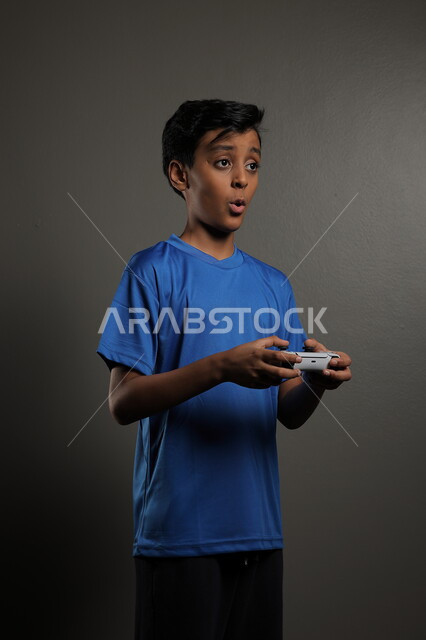 قضاء اوقات ممتعة باللعب في المنزل، استخدام التقنيات الحديثة للتسلية والترفيه، بورتريه لولد عربي خليجي سعودي يرتدي الزي الكاجوال يقوم باللعب في ألعاب الفيديو بايماءات الاندماج والتركيز، خلفية رمادية