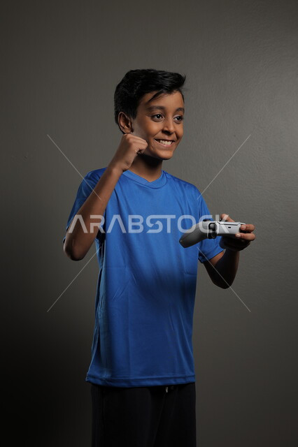 قضاء اوقات ممتعة باللعب في المنزل، استخدام التقنيات الحديثة للتسلية والترفيه، بورتريه لولد عربي خليجي سعودي يرتدي الزي الكاجوال يقوم باللعب في ألعاب الفيديو بايماءات الاندماج والتركيز، خلفية رمادية