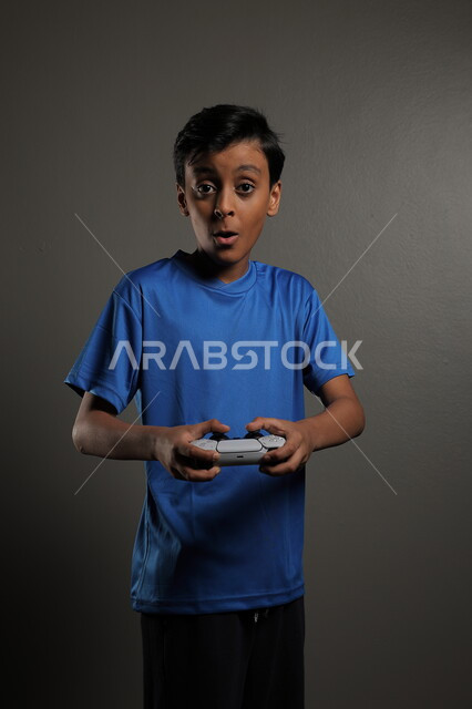 قضاء اوقات ممتعة باللعب في المنزل، استخدام التقنيات الحديثة للتسلية والترفيه، بورتريه لولد عربي خليجي سعودي يرتدي الزي الكاجوال يقوم باللعب في ألعاب الفيديو بايماءات الاندماج والتركيز، خلفية رمادية