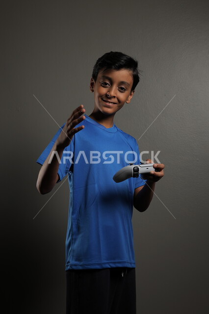  استخدام التقنيات الحديثة للتسلية والترفيه، قضاء اوقات ممتعة باللعب في المنزل، بورتريه لولد عربي خليجي سعودي يرتدي الزي الكاجوال يقوم باللعب في ألعاب الفيديو بايماءات الاندماج والتركيز، خلفية رمادية