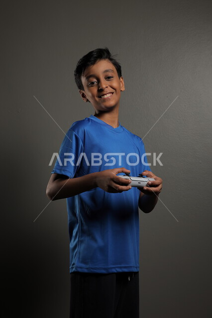 Using modern technologies for entertainment and fun, spending fun times playing at home, portrait of a Saudi Arabian Gulf boy wearing casual clothes playing video games with gestures of integration and focus, gray background