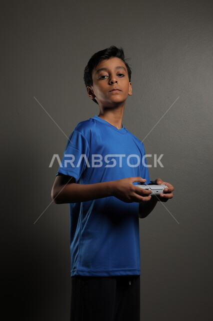  استخدام التقنيات الحديثة للتسلية والترفيه، قضاء اوقات ممتعة باللعب في المنزل، بورتريه لولد عربي خليجي سعودي يرتدي الزي الكاجوال يقوم باللعب في ألعاب الفيديو بايماءات الاندماج والتركيز، خلفية رمادية