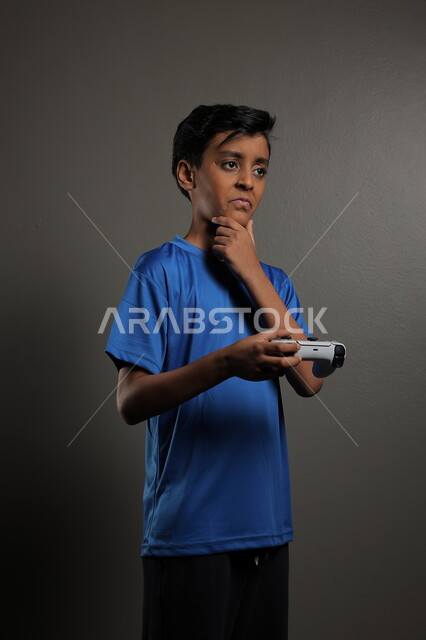  استخدام التقنيات الحديثة للتسلية والترفيه، قضاء اوقات ممتعة باللعب في المنزل، بورتريه لولد عربي خليجي سعودي يرتدي الزي الكاجوال يقوم باللعب في ألعاب الفيديو بايماءات الاندماج والتركيز، خلفية رمادية