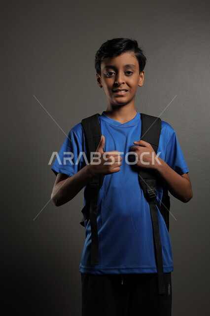 الوقوف باستقامة بايماءات السعادة للعودة إلى المدارس ,  بورتريه لطالب عربي خليجي سعودي يرتدي الزي الكاجوال و يحمل الحقيبة المدرسية وينظر إلى الكاميرا ، الإستعداد للعام  الجديد، الشعور بالحماس والنشاط ببداية السنة الدراسية ، خلفية رمادية