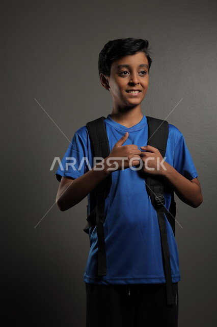 الوقوف باستقامة بايماءات السعادة للعودة إلى المدارس ,  بورتريه لطالب عربي خليجي سعودي يرتدي الزي الكاجوال و يحمل الحقيبة المدرسية وينظر إلى الكاميرا ، الإستعداد للعام  الجديد، الشعور بالحماس والنشاط ببداية السنة الدراسية ، خلفية رمادية