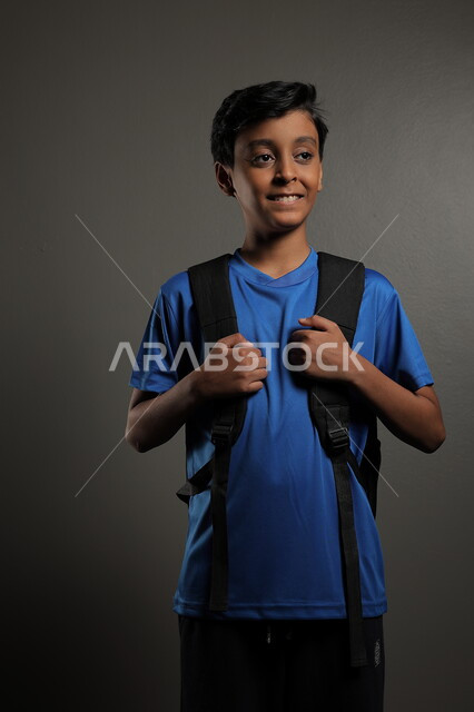 الوقوف باستقامة بايماءات السعادة للعودة إلى المدارس ,  بورتريه لطالب عربي خليجي سعودي يرتدي الزي الكاجوال و يحمل الحقيبة المدرسية وينظر إلى الكاميرا ، الإستعداد للعام  الجديد، الشعور بالحماس والنشاط ببداية السنة الدراسية ، خلفية رمادية