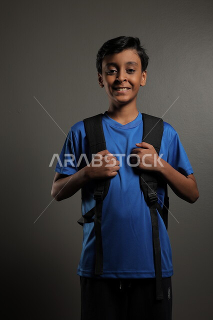 الوقوف باستقامة بايماءات السعادة للعودة إلى المدارس ,  بورتريه لطالب عربي خليجي سعودي يرتدي الزي الكاجوال و يحمل الحقيبة المدرسية وينظر إلى الكاميرا ، الإستعداد للعام  الجديد، الشعور بالحماس والنشاط ببداية السنة الدراسية ، خلفية رمادية