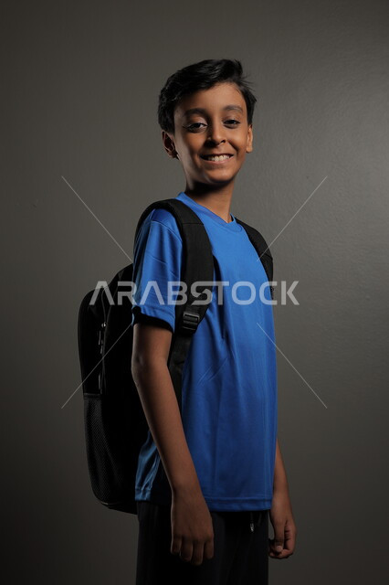 الوقوف باستقامة بايماءات السعادة للعودة إلى المدارس ,  بورتريه لطالب عربي خليجي سعودي يرتدي الزي الكاجوال و يحمل الحقيبة المدرسية وينظر إلى الكاميرا ، الإستعداد للعام  الجديد، الشعور بالحماس والنشاط ببداية السنة الدراسية ، خلفية رمادية
