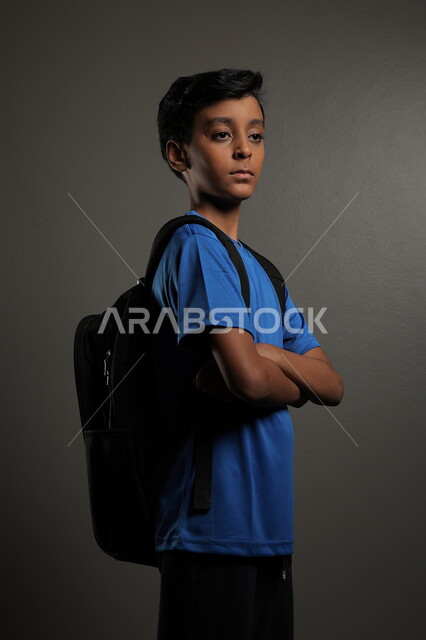 الوقوف باستقامة بايماءات السعادة للعودة إلى المدارس ,  بورتريه لطالب عربي خليجي سعودي يرتدي الزي الكاجوال و يحمل الحقيبة المدرسية وينظر إلى الكاميرا ، الإستعداد للعام  الجديد، الشعور بالحماس والنشاط ببداية السنة الدراسية ، خلفية رمادية