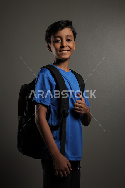 الوقوف باستقامة بايماءات السعادة للعودة إلى المدارس ,  بورتريه لطالب عربي خليجي سعودي يرتدي الزي الكاجوال و يحمل الحقيبة المدرسية وينظر إلى الكاميرا ، الإستعداد للعام  الجديد، الشعور بالحماس والنشاط ببداية السنة الدراسية ، خلفية رمادية