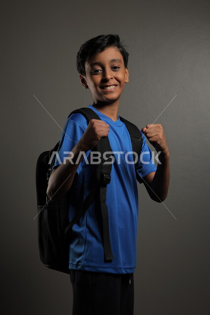 الإستعداد للعام  الجديد، الوقوف باستقامة بايماءات السعادة للعودة إلى المدارس ,  بورتريه لطالب عربي خليجي سعودي يرتدي الزي الكاجوال و يحمل الحقيبة المدرسية وينظر إلى الكاميرا ، الشعور بالحماس والنشاط ببداية السنة الدراسية ، خلفية رمادية