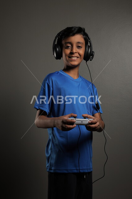 مفهوم المتعة والتسلية، قضاء وقت ممتع في المنزل، أنشطة ترفيهية شبابية ممتعه ,الاستمتاع بالتقنيات والتكنولوجيا الحديثة, بورتريه لشاب عربي خليجي سعودي يرتدي سماعة الرأس يقوم باللعب بإيماءات السرور والسعادة، خلفية رمادية