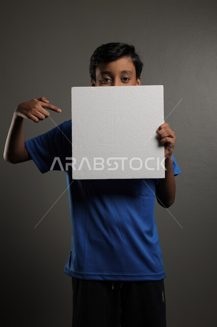 استخدام لوحات توضيحية، مفهوم الإعلان والتسويق لمكان ما، الترويج للخدمات والمنتجات، موك اب مربع صغير، بورتريه لولد عربي خليجي سعودي يرتدي الزي الكاجوال يمسك بيده لوحة بيضاء فارغة،  لوح إعلاني فارغ ، خلفية رمادية