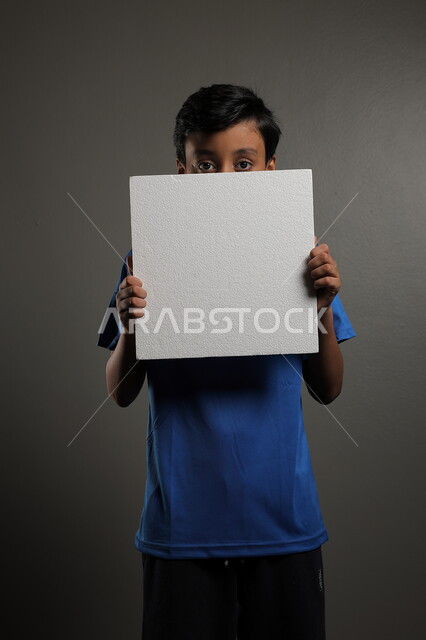 استخدام لوحات توضيحية، مفهوم الإعلان والتسويق لمكان ما، الترويج للخدمات والمنتجات، موك اب مربع صغير، بورتريه لولد عربي خليجي سعودي يرتدي الزي الكاجوال يمسك بيده لوحة بيضاء فارغة،  لوح إعلاني فارغ ، خلفية رمادية