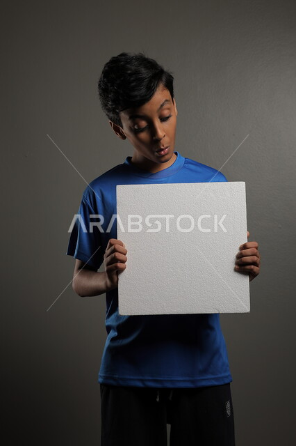استخدام لوحات توضيحية، مفهوم الإعلان والتسويق لمكان ما، الترويج للخدمات والمنتجات، موك اب مربع صغير، بورتريه لولد عربي خليجي سعودي يرتدي الزي الكاجوال يمسك بيده لوحة بيضاء فارغة،  لوح إعلاني فارغ ، خلفية رمادية