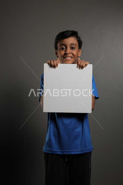  الترويج للخدمات والمنتجات، استخدام لوحات توضيحية، مفهوم الإعلان والتسويق لمكان ما، موك اب مربع صغير، بورتريه لولد عربي خليجي سعودي يرتدي الزي الكاجوال يمسك بيده لوحة بيضاء فارغة،  لوح إعلاني فارغ ، خلفية رمادية
