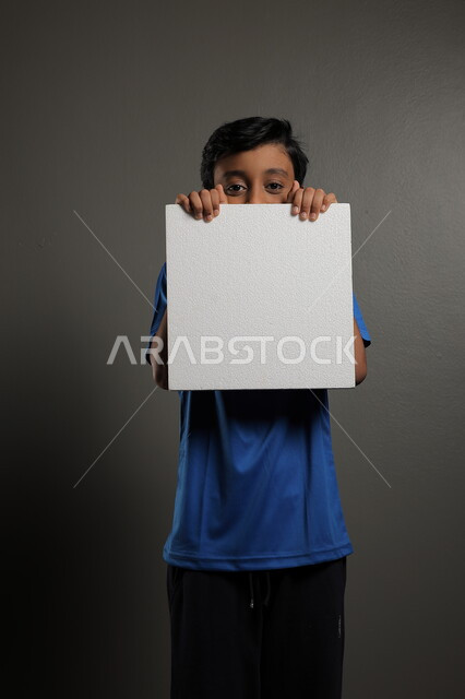  الترويج للخدمات والمنتجات، استخدام لوحات توضيحية، مفهوم الإعلان والتسويق لمكان ما، موك اب مربع صغير، بورتريه لولد عربي خليجي سعودي يرتدي الزي الكاجوال يمسك بيده لوحة بيضاء فارغة،  لوح إعلاني فارغ ، خلفية رمادية