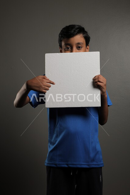  الترويج للخدمات والمنتجات، استخدام لوحات توضيحية، مفهوم الإعلان والتسويق لمكان ما، موك اب مربع صغير، بورتريه لولد عربي خليجي سعودي يرتدي الزي الكاجوال يمسك بيده لوحة بيضاء فارغة،  لوح إعلاني فارغ ، خلفية رمادية
