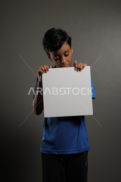  الترويج للخدمات والمنتجات، استخدام لوحات توضيحية، مفهوم الإعلان والتسويق لمكان ما، موك اب مربع صغير، بورتريه لولد عربي خليجي سعودي يرتدي الزي الكاجوال يمسك بيده لوحة بيضاء فارغة،  لوح إعلاني فارغ ، خلفية رمادية