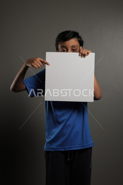 الترويج للخدمات والمنتجات، استخدام لوحات توضيحية، مفهوم الإعلان والتسويق لمكان ما، موك اب مربع صغير، بورتريه لولد عربي خليجي سعودي يرتدي الزي الكاجوال يمسك بيده لوحة بيضاء فارغة،  لوح إعلاني فارغ ، خلفية رمادية
