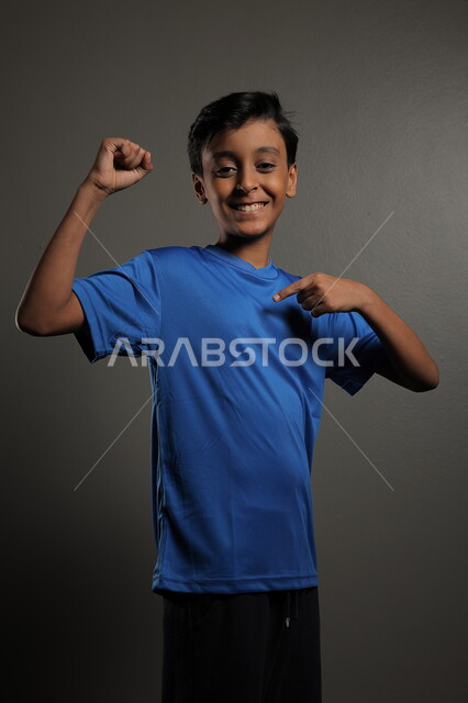 النظر إلى مكان ما، ايماءات البشاشة والبراءة، الاهتمام بالمظهر الخارجي للأولاد , بورتريه مقرب لولد عربي خليجي سعودي مبتسم يرتدي الزي الكاجوال يقف باستقامة، خلفية رمادية