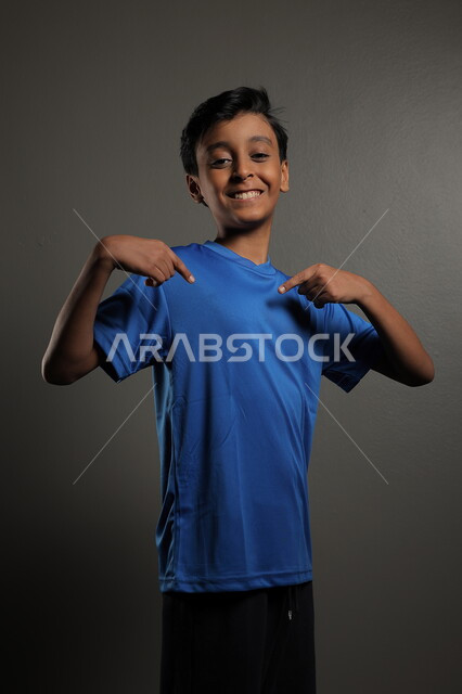 النظر إلى مكان ما، ايماءات البشاشة والبراءة، الاهتمام بالمظهر الخارجي للأولاد , بورتريه مقرب لولد عربي خليجي سعودي مبتسم يرتدي الزي الكاجوال يقف باستقامة، خلفية رمادية