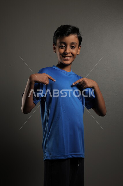 النظر إلى مكان ما، ايماءات البشاشة والبراءة، الاهتمام بالمظهر الخارجي للأولاد , بورتريه مقرب لولد عربي خليجي سعودي مبتسم يرتدي الزي الكاجوال يقف باستقامة، خلفية رمادية