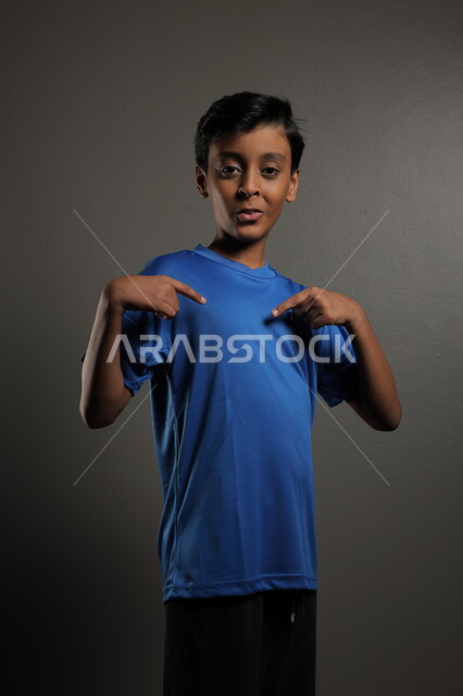 النظر إلى مكان ما، ايماءات البشاشة والبراءة، الاهتمام بالمظهر الخارجي للأولاد , بورتريه مقرب لولد عربي خليجي سعودي مبتسم يرتدي الزي الكاجوال يقف باستقامة، خلفية رمادية