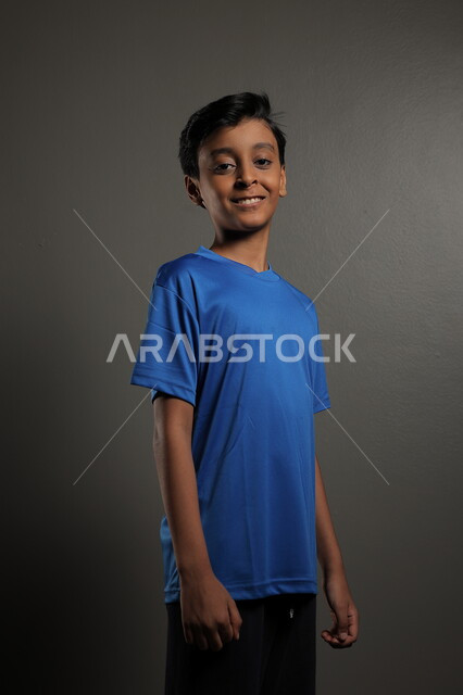 النظر إلى مكان ما، ايماءات البشاشة والبراءة، الاهتمام بالمظهر الخارجي للأولاد , بورتريه مقرب لولد عربي خليجي سعودي مبتسم يرتدي الزي الكاجوال يقف باستقامة، خلفية رمادية