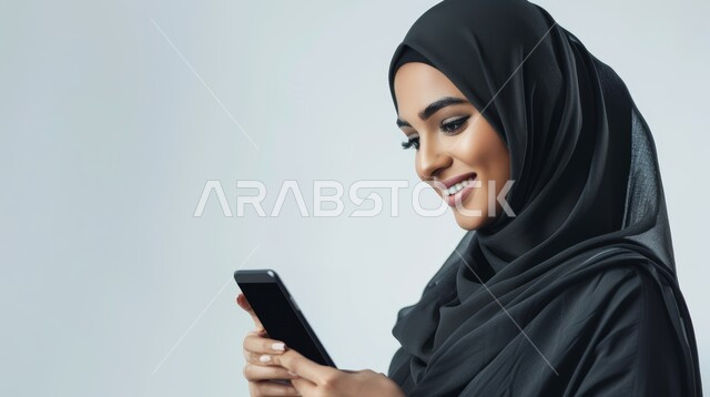 إيماءات السعادة والسرور، استخدام الاجهزة التقنية الحديثة، بورتريه مقرب لامرأة عربية خليجية سعودية محجبة تحمل بيدها الهاتف المحمول تقوم بتصفح وسائل التواصل الاجتماعي، الدردشة مع الأهل والأصدقاء، خلفية رمادية