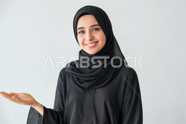 مفهوم البشاشة والترحيب والاستقبال، إيماءات السعادة والسرور، بورتريه مقرب لامرأة عربية خليجية سعودية محجبة تنظر الى الكاميرا ترفع يدها للأعلى بتعابير حمل شيء ما، رفع عنصر هولوجرامي، خلفية رمادية