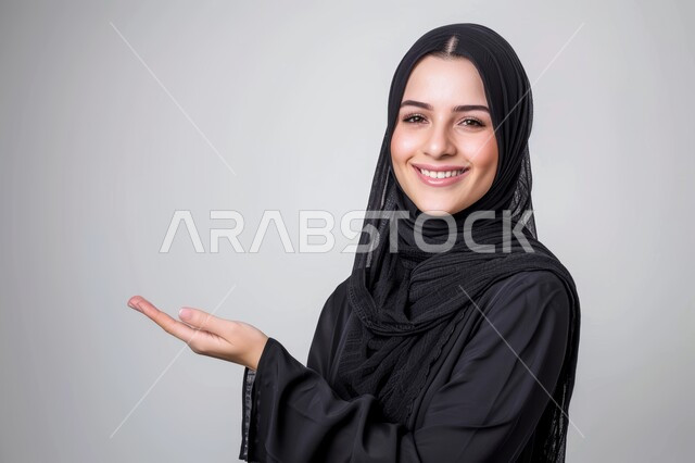مفهوم البشاشة والترحيب والاستقبال، إيماءات السعادة والسرور، بورتريه مقرب لامرأة عربية خليجية سعودية محجبة تنظر الى الكاميرا ترفع يدها للأعلى بتعابير حمل شيء ما، رفع عنصر هولوجرامي، خلفية رمادية