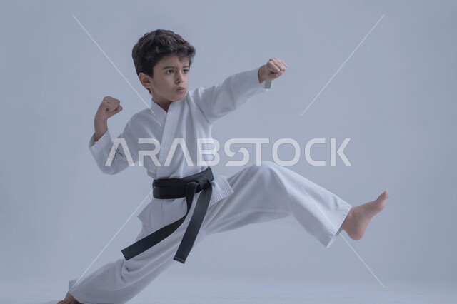 تطبيق حركات الكاتا, بورتريه لطفل عربي خليجي سعودي يمارس رياضة الكاراتيه اليابانية, زي الكاراتيه الأبيض مع الحزام الأسود، فنون الدفاع عن النفس، التدرب على حركات قتالية خاصة، صورة الجسم بالطول الكامل, خلفية رمادية