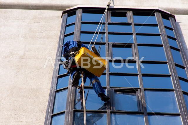 المنشآت الحديثة بديكورات وتصاميم متميزة في الإمارات العربية المتحدة، الفن الهندسي المعماري للأبراج وناطحات السحاب، عامل عربي خليجي إماراتي يقوم بتنظيف الزجاج الخارجي لبناء مرتفع شاهق، خدمة التنظيف في العمارات المرتفعة