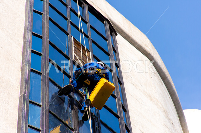 المنشآت الحديثة بديكورات وتصاميم متميزة في الإمارات العربية المتحدة، الفن الهندسي المعماري للأبراج وناطحات السحاب، عامل عربي خليجي إماراتي يقوم بتنظيف الزجاج الخارجي لبناء مرتفع شاهق، خدمة التنظيف في العمارات المرتفعة