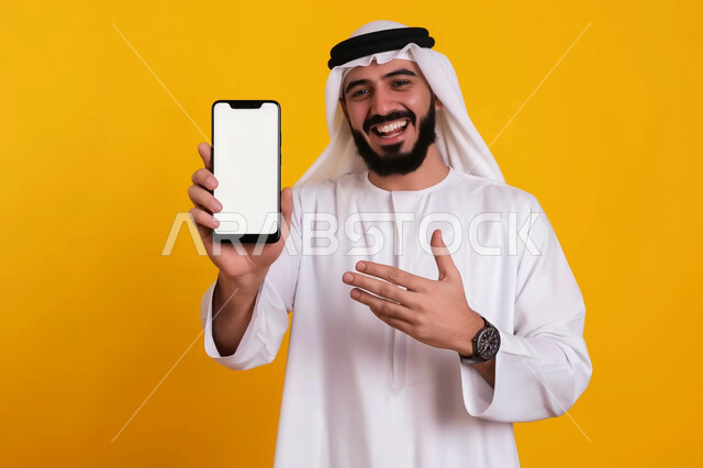 Using modern technology devices in daily life, looking at the camera with gestures of happiness and pleasure, close-up portrait of a young Saudi Arabian Gulf man wearing traditional thobe and shemagh holding a mobile phone and pointing at it with his hand, displaying a blank white screen via mobile, yellow background