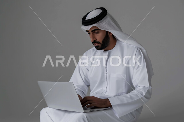 إدارة شؤون الأعمال وتنظيمها عبر الاجهزة التقنية، دمج العمل مع التقنية والتكنولوجيا، بورتريه مقرب لرجل اعمال عربي خليجي سعودي يرتدي الزي التقليدي والغترة يعمل على جهاز الحاسوب المحمول بإيماءات التركيز، مهن ووظائف ادارية مكتبية، خلفية رمادية