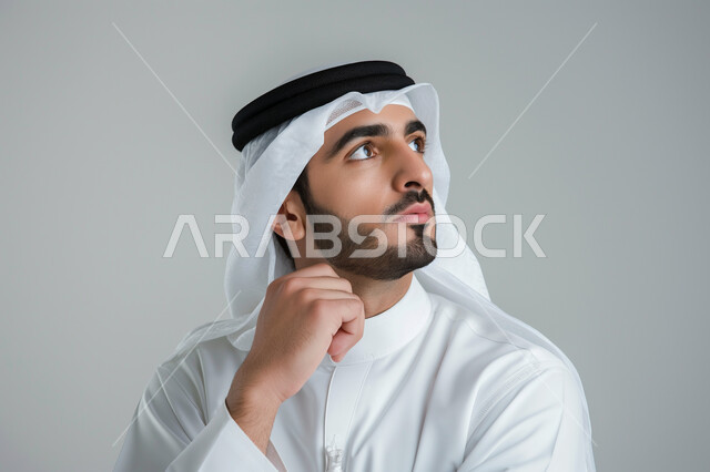 Gestures of confusion and anxiety, trying to find a solution to a problem, contemplation, concentration and planning for the future, close-up portrait of a Saudi Arabian Gulf man wearing traditional thobe and looking away with an expression of thought and contemplation, looking absentmindedly at a place, gray background