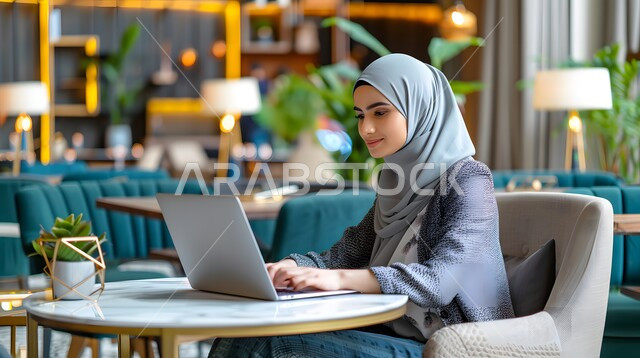 دمج التقنيات المتطورة في الحياة اليومية, استخدام الأجهزة الحديثة لإنجاز المهام, شابة عربية خليجية سعودية ترتدي الحجاب تجلس على الطاولة و تعمل على جهاز الحاسوب المحمول