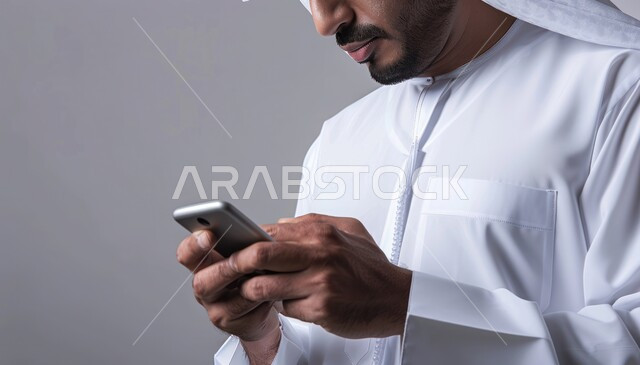  تصفح مواقع الانترنت، إرسال رسالة نصية عبر الجوال, بورتريه مقرب ليدي رجل عربي خليجي سعودي يرتدي الثوب التقليدي يقوم بمتابعة آخر أخبار السوشال ميديا عبر الهاتف المحمول, التواصل مع الأصدقاء والأقارب ،دمج التكنولوجيا الحديثة بالحياة العملية، خلفية  رمادية