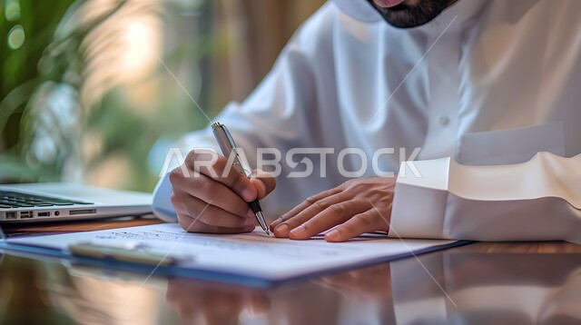 مهن ووظائف مكتبيه , انجاز المهام وتحقيق الاهداف, ملفات ووثائق خاصة بالعمل, صورة مقربة ليد رجل عربي خليجي سعودي يرتدي الثوب التقليدي والشماغ يقوم بتدوين وكتابة الملاحظات على الورق , متابعه اعمال الشركه ودراسه وتتبع المشاريع