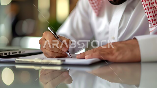  متابعه اعمال الشركه ودراسه وتتبع المشاريع , انجاز المهام وتحقيق الاهداف ,  مهن ووظائف مكتبيه , ملفات ووثائق خاصة بالعمل, صورة مقربة ليد رجل عربي خليجي سعودي يرتدي الثوب التقليدي والشماغ يقوم بتدوين وكتابة الملاحظات على الورق 