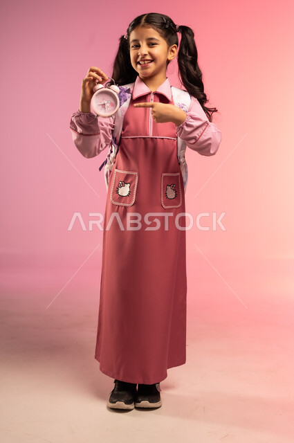 مفهوم تنظيم و إدارة الوقت, الاستعداد للعام الدراسي الجديد, التعليم في المملكة العربية السعودية, بورتريه لفتاة عربية خليجية سعودية ترتدي الزي المدرسي و تحمل على ظهرها حقيبة تمسك بيدها ساعة المنبه و تشير إليها بإصبع السبابة, الجسم بالطول الكامل, خلفية ملونة