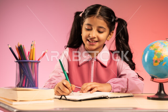 مفهوم العودة إلى المدارس, التعليم في المملكة العربية السعودية, الاستعداد للعام الدراسي الجديد, بورتريه مقرب لفتاة عربية خليجية سعودية ترتدي الزي المدرسي تجلس على الطاولة و تقوم بالكتابة على الدفتر, خلفية ملونة
