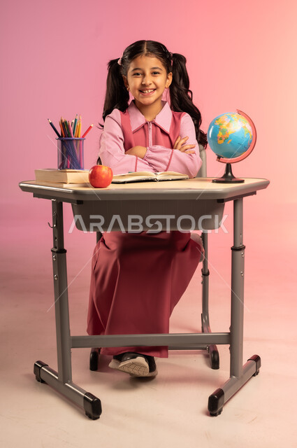 البهجة بالعودة إلى المدارس, الاستعداد للعام الدراسي الجديد, التعليم في المملكة العربية السعودية, بورتريه لفتاة عربية خليجية سعودية ترتدي الزي المدرسي تجلس على الطاولة و تنظر إلى الكاميرا بإيماءات السعادة و السرور, الجسم بالطول الكامل, خلفية ملونة