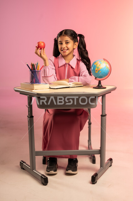 النظر إلى الكاميرا بإيماءات السعادة و السرور, الاستعداد للعام الدراسي الجديد, التعليم في المملكة العربية السعودية, بورتريه لفتاة عربية خليجية سعودية ترتدي الزي المدرسي تجلس على الطاولة تمسك بيدها تفاحة حمراء, الجسم بالطول الكامل, خلفية ملونة