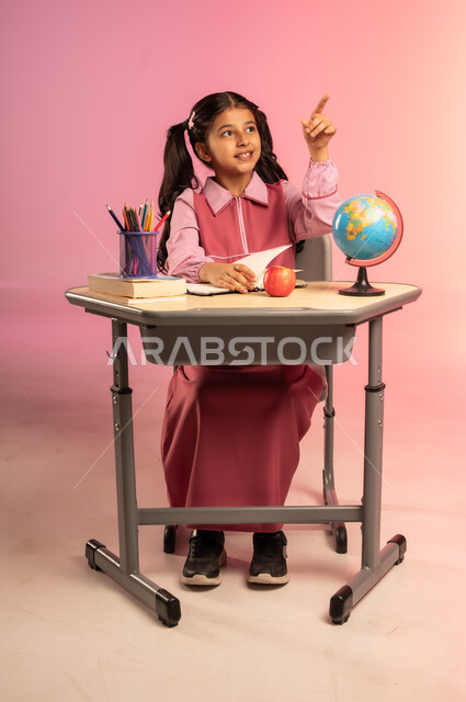 الاستعداد للعام الدراسي الجديد, مفهوم العودة إلى المدارس, التعليم في المملكة العربية السعودية, بورتريه لفتاة عربية خليجية سعودية ترتدي الزي المدرسي تجلس على الطاولة و تشير بإصبع السبابة إلى الأعلى, الجسم بالطول الكامل, خلفية ملونة