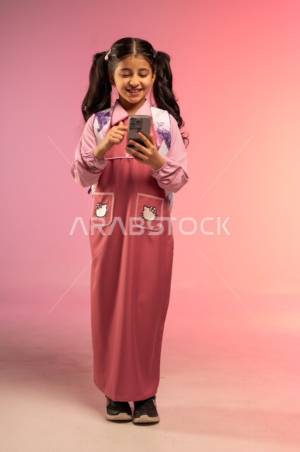 استخدام الأجهزة التقنية الحديثة في الحياة اليومية, الاستعداد للعام الدراسي الجديد, مفهوم العودة إلى المدارس, بورتريه لفتاة عربية خليجية سعودية ترتدي الزي المدرسي تقوم باستعمال الهاتف المحمول, الجسم بالطول الكامل, خلفية ملونة