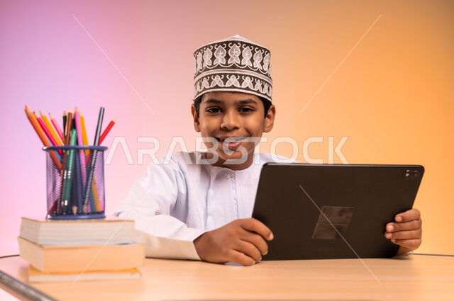 طرائق تعليمية جديدة متطورة، بورتريه لطالب عربي خليجي عماني يرتدي الثوب التقليدي والقبعة العمانية يجلس في مقعده ويمسك بالجهاز اللوحي، النظر إلى الكاميرا بإيماءات السعادة والسرور، موسم العودة إلى المدارس، دمج التكنولوجيا بالتعليم في المدارس الحديثة، خلفية ملونة