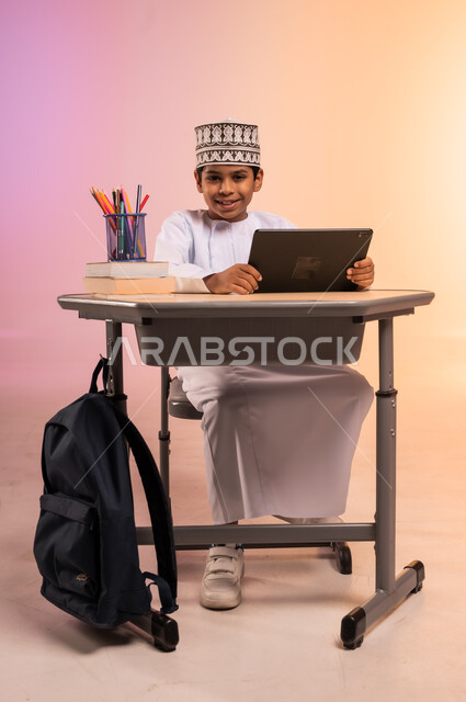 طرائق تعليمية جديدة متطورة، بورتريه لطالب عربي خليجي عماني يرتدي الثوب التقليدي والقبعة العمانية يجلس في مقعده ويمسك بالجهاز اللوحي، النظر إلى الكاميرا بإيماءات السعادة والسرور، موسم العودة إلى المدارس، دمج التكنولوجيا بالتعليم في المدارس الحديثة، خلفية ملونة
